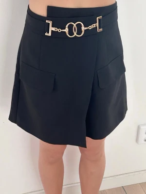 Svarta skorts med gulddetaljer - Trendiga svarta skorts med snygg guldfärgad kedjedetalj framtill. Skortsen har en stilren design med omlottkänsla och är perfekt för dig som vill ha både kjol-look och shortskomfort. Nya, endast provade. Storlek XS-S.