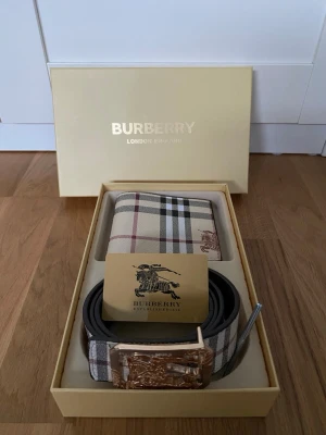 B plånbok och bälte beige rutigt mönster - Snygg plånbok och bälte från B i klassiskt beige rutigt mönster med bruna, svarta och röda detaljer. Plånboken har Burberrys ikoniska riddarlogga tryckt på framsidan och är tillverkad i tåligt material. Perfekt accessoar för dig som gillar stilrena designer. Man kan även vända sida på bältet till svart. 