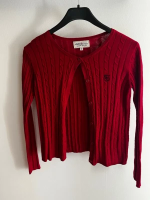 Röd kabelstickad kofta Hampton Republic - Snygg röd kabelstickad kofta från Hampton Republic med rund halsringning och knappar framtill. Liten broderad logga på bröstet. Långärmad och i mjukt material som passar perfekt till lager på lager. Aldrig använd.
