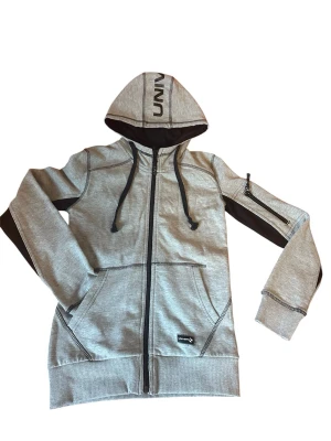 Grå zip-hoodie från Universe - Snygg grå hoodie med dragkedja från Universe. Svarta detaljer på ärmar och huva, samt svart snörning. Praktiska fickor fram och en extra ficka med dragkedja på ärmen. Mjuk bomullsblandning och ribbade muddar för skön passform.