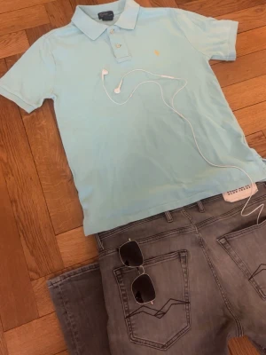 Ljusblå pikétröja från Polo Ralph Lauren - Snygg ljusblå pikétröja från Polo Ralph Lauren med klassisk krage och två knappar framtill. Tröjan har korta ärmar och den ikoniska broderade loggan i gult på bröstet. Perfekt till jeans eller shorts för en clean och fräsch look.