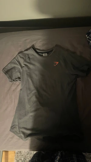 Svart Gymshark t-shirt - Snygg svart t-shirt från Gymshark med korta ärmar och orange logga på bröstet. På ryggen finns en stor orange detalj med texten GYMSHARK. Perfekt för dig som gillar sportig och stilren design.