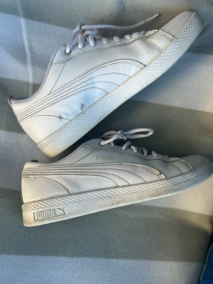 Vita Puma sneakers i läder - Säljer ett par klassiska vita sneakers från Puma i läder med lågt skaft och vita snören. Skorna har diskret Puma-logga på sidan och hälen samt en stilren, enkel design som passar till det mesta. Yttersulan är vit med mönstrad kant.