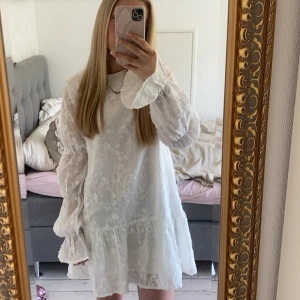 Vit blommig klänning från NA-KD❤️ - Superfin vit kortklänning med blommigt broderi och volang nertill. Klänningen har långa, lätt puffiga ärmar med volang vid ärmslut och rund halsringning. Perfekt för sommarens alla tillfällen och ger en romantisk vibe.