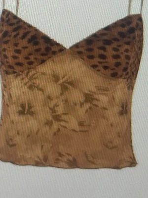 Leopardmönstrad linnetopp - Trendig linnetopp med leopardmönstrad byst och tunna axelband. Toppen har en beige botten med subtilt blommönster och är tillverkad i ett lätt, glansigt material som ger en lyxig känsla. Perfekt för dig som vill sticka ut med en unik och djärv look.