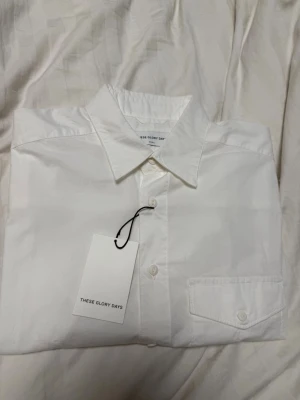 Vit skjorta från These Glory Days - Klassisk vit skjorta från These Glory Days med krage och bröstficka. Skjortan har knäppning framtill och är tillverkad i bomull, perfekt för en clean och stilren look. Nypris: 799kr.
