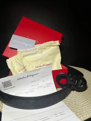 Svart skärp från Salvatore Ferragamo - Snyggt svart bälte från Salvatore Ferragamo med ikoniskt dubbel-Gancio-spänne i matt finish. Bältet har präglat mönster längs hela remmen och levereras med originalförpackning och dustbag. Perfekt accessoar för att lyfta din outfit.