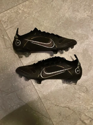 Nike Mercurial vapor elite  fotbollsskor - Säljer ett par svarta Nike Mercurial fotbollsskor med gulddetaljer och vita loggor. Skorna har låg profil, snörning och dobbar för spel på gräs. Tillverkade i syntetmaterial för bra grepp och kontroll på planen. Snygg design med moderna detaljer och stilrena linjer.
