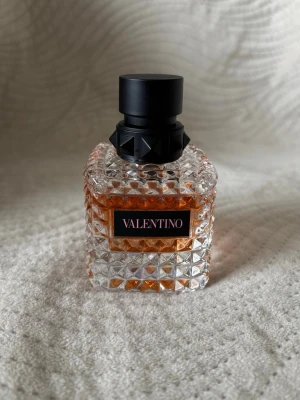 Valentino parfym i lyxig glasflaska 50ml - Exklusiv parfym från Valentino i en fyrkantig, transparent glasflaska med nitar och mönstrad yta. Flaskan har en svart, matt kork med dekorativa detaljer och innehållet är bärnstensfärgat. Perfekt för dig som gillar eleganta och unika dofter.