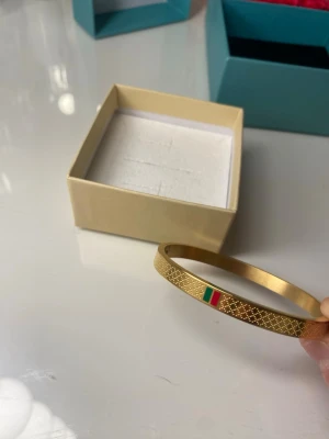 18K Guld pläterad Armband  - Snyggt armband i guldfärgad metall med graverat rutmönster och en liten detalj. Armbandet är stelt och har en modern, minimalistisk design som sticker ut.