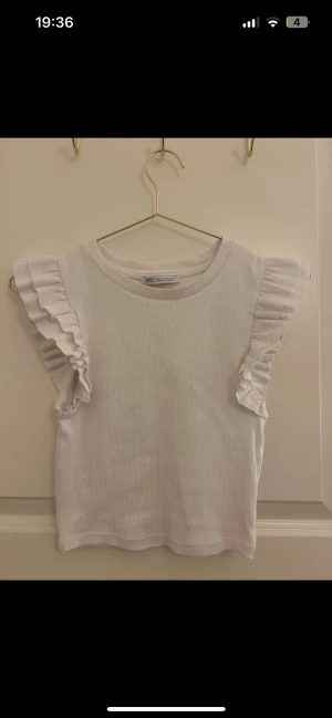 Vit ribbad topp med volangärmar från Zara - Supersöt vit ribbad topp från Zara med volangdetaljer på ärmarna. Ärmlös modell med rund halsringning och stretchigt material som sitter snyggt. Perfekt för dig som gillar en clean och feminin look.