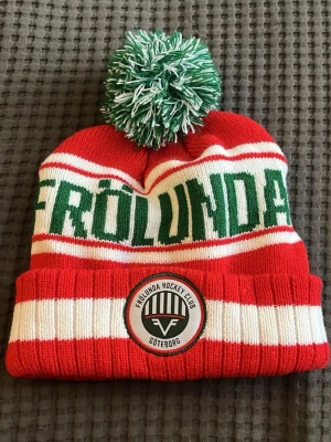 Frölunda HC röd-vit mössa med tofs - Ny utan prislapp, aldrig använd. Stickad mössa i rött, vitt och grönt med stor grön-vit tofs på toppen. Frölunda står i grönt över mössan och klubbens logga sitter framtill på uppvikt kant. Perfekt för dig som vill visa ditt stöd för Frölunda HC.