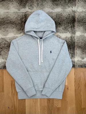 Ljusgrå Polo Ralph Lauren Fleece Hoodie - Storlek S - Hej! 👋 Säljer en klassisk ljusgrå (grey heather) Polo Ralph Lauren hoodie i storlek S (Herr). Den har den ikoniska mörkblå pony-loggan broderad på bröstet 🐎 Tröjan är i en skön, kraftig bomullsblandning med mjuk fleece-insida ✨ Mycket gott skick utan fläckar eller hål! Sitter bra på mig (177cm) och passar perfekt som basplagg året om 👕 Denna modell saknar QR-kod, men alla invändiga tvättlappar sitter kvar och garanterar äktheten ✅ Fraktar snabbt! 🚛📦💬💯 