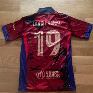 FC Barcelona Lamine Yamal tröja Nike - Säljer ett FC Barcelona sett med Lamine Yamal och nummer 19 på ryggen. Tröjan är röd med blå detaljer, har coola grafiska mönster och klubbmärke på bröstet. Korta ärmar, Dri-FIT material och sponsorer som Spotify och UNHCR tryckta på. Perfekt för alla Barca-fans!