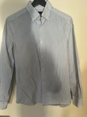 Randig skjorta från Riley slim fit - Klassisk vit skjorta med tunna blå ränder från Riley. Slim fit-modell med krage och knäppning framtill. Perfekt för dig som gillar en stilren och tidlös look. Tillverkad i ett mjukt och lätt bomullstyg.