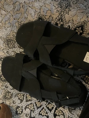 Svarta sandaler med breda remmar - Säljer ett par svarta sandaler med breda korsade remmar och justerbart spänne vid ankeln. Sandalerna har en stilren och enkel design med öppen tå och häl. Materialet ser ut att vara textil eller syntet. Perfekta för sommaren och lätta att matcha med olika outfits.