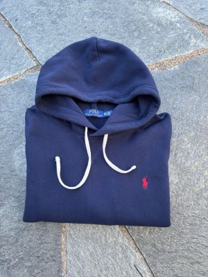 Mörkblå hoodie från Polo Ralph Lauren - Snygg mörkblå hoodie från Polo Ralph Lauren med vit dragsko och den klassiska röda loggan broderad på bröstet. Hoodien har en avslappnad passform och är tillverkad i mjukt bomullsmaterial. Perfekt för en chill och stilren look.