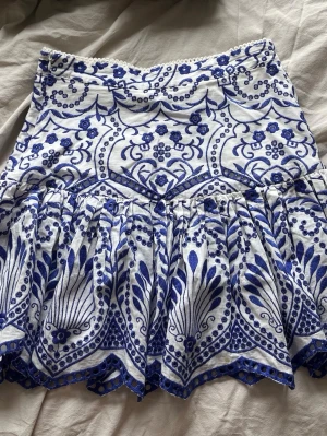 Vit och blå broderad kortkjol - Supersöt kortkjol i vitt med blått broderat mönster och dekorativa knappar bak. Kjolen har volangkant och ett unikt, detaljerat mönster som ger en bohemisk vibe. Perfekt för dig som vill sticka ut med en färgglad och somrig look.