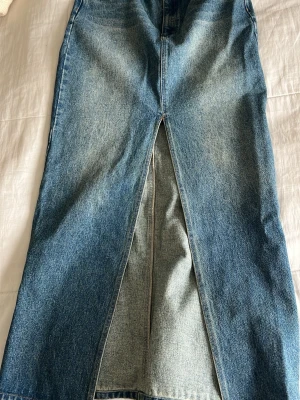Lång jeanskjol med slits från Icons - Säljer en blå lång jeanskjol från Icons i klassisk denim med en snygg slits framtill. Kjolen har hög midja och raka linjer, perfekt till sneakers eller boots. Enkel att styla och ett måste i garderoben!