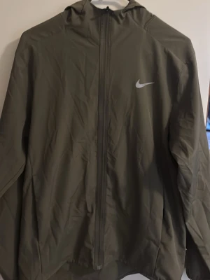 Olivgrön Nike vindjacka med huva - Stilren olivgrön vindjacka från Nike med hel dragkedja framtill och huva. Jackan har en minimalistisk design med Nike-logga på bröstet och elastiska muddar vid ärmsluten. Tillverkad i lätt, vindtätt material som passar perfekt för blåsiga dagar. Seriöst använt 2 gånger!