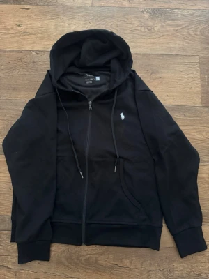 Svart hoodie från Polo Ralph Lauren - Klassisk svart hoodie med dragkedja från Polo Ralph Lauren. Hoodien har huva med dragsko, två fickor framtill och den ikoniska vita logon broderad på bröstet. Tillverkad i mjukt bomullsmaterial och har ribbade muddar vid ärmslut och nederkant.