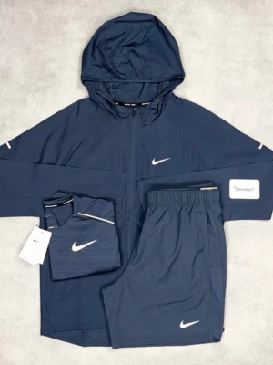 Mörkblå Nike träningsset med hoodie - NIKE MILLER Snyggt mörkblått träningsset från Nike med matchande hoodie, shorts och t-shirt. Hoodien har dragkedja, huva och vita Nike-loggor på bröst och ärmar. Shortsen har resår i midjan och t-shirten har diskret randigt mönster. Perfekt för träning eller chill.