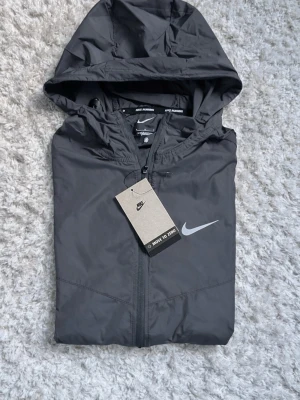 Svart Nike vindjacka med huva - Snygg svart vindjacka från Nike med huva och dragkedja framtill, prislappen sitter kvar. Storlek S. Fraktar inom 24 timmar 
