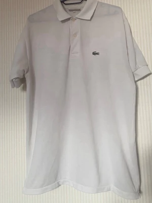 Vit pikétröja från Lacoste - Klassisk vit pikétröja från Lacoste med korta ärmar och krage. Tröjan har två knappar framtill och den ikoniska gröna krokodilloggan på bröstet. Tillverkad i mjuk bomull som känns skön mot huden. Perfekt till en avslappnad och stilren look.