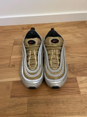 Nike Air Max 97 Silver/Gold sneakers - Nike Air Max 97 sneakers i silver och guld med svarta och röda detaljer. Skorna har den ikoniska vågiga designen, synlig Air-sula och mesh- samt syntetmaterial. Swoosh-logga på sidan och plös. Perfekta för dig som gillar streetwear och sportig stil. Dom har reflexer.