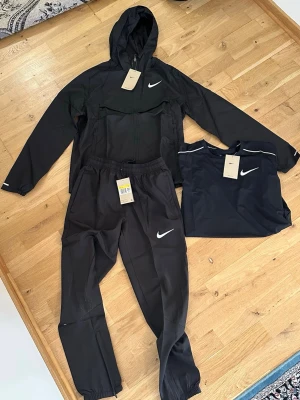 Svart Nike träningsset med hoodie - Säljer ett svart träningsset från Nike med hoodie, byxor och en matchande t-shirt. Setet har vita Nike-loggor och är gjort i ett lätt, funktionellt material som passar perfekt till träning eller sport. Jackan har dragkedja och huva, byxorna har resår i midjan och benslut.