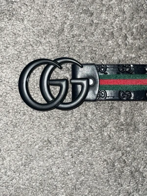  svart bälte med GG-spänne - Snyggt svart bälte från  med klassiskt GG-spänne i matt svart metall. Bältet har präglade GG-loggor längs hela remmen och en ikonisk grön och röd rand i mitten. Perfekt accessoar för att lyfta din outfit.