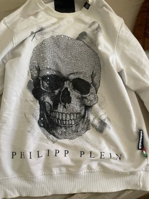 Vit hoodie från Philipp Plein - Säljer en vit hoodie från Philipp Plein med tryck framtill och märkets logga broderad längst ner. Hoodien har ribbade muddar och en klassisk passform. Materialet känns mjukt och skönt, perfekt för dig som gillar streetwear och exklusiva märken.