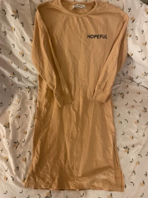 Beige långärmad klänning HOPEFUL barn st 134/140 - Beige långärmad klänning från i dig denim med texten 'HOPEFUL' tryckt på bröstet. Klänningen har en rak passform, rund halsringning och en slits på sidan för extra rörelsefrihet. Tillverkad i mjuk bomull. Barn storlek 134/140