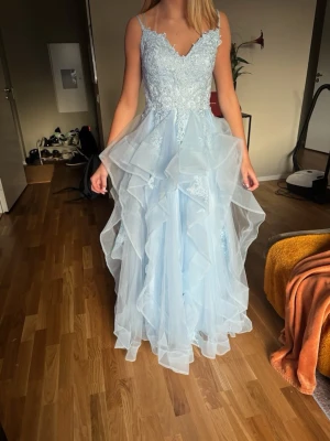 Ljusblå långklänning med spets och volang - Drömmig ljusblå långklänning med tunna axelband, spetsdetaljer upptill och flera lager volangig tyll i kjolen. Klänningen har v-ringning och en elegant, flowy siluett som ger en sagolik känsla. Perfekt för bal eller speciella tillfällen.