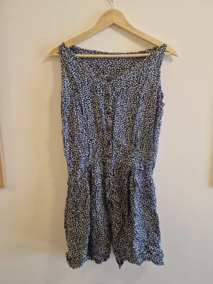Blåvit prickig ärmlös jumpsuit - Säljer en blå ärmlös jumpsuit med vita prickar och knappar framtill. Modellen har en avslappnad passform och är gjord i ett lätt, mjukt tyg som känns skönt mot huden. Perfekt för dig som gillar en lekfull och bekväm stil.
