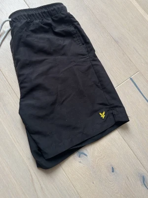 Svarta badbyxor från Lyle & Scott - Snygga svarta badbyxor från Lyle & Scott med gul logga broderad på benet. Klassisk modell med resår i midjan och vit mesh på insidan för extra komfort. Perfekta för stranden eller poolen.