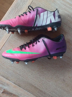 Nike Mercurial rosa fotbollsskor - Säljer ett par Nike Mercurial fotbollsskor i en snygg rosa färg med gröna och svarta detaljer. Skorna har klassisk låg profil, snörning och dobbar för spel på gräs. Tillverkade i syntetmaterial för lätt känsla och bra bollkontroll. Perfekta för dig som vill sticka ut på planen. Kan har lite slitage inne i skorna vid hälen men annars är dom bra för att var 13 år gammla.