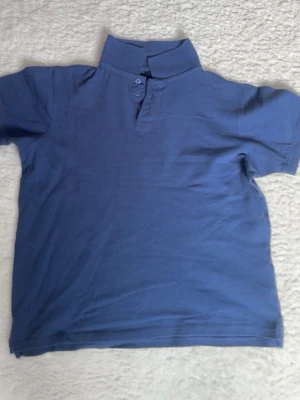 Blå piké t-shirt med knappar - Säljer en stilren blå piké t-shirt med korta ärmar och klassisk krage. Tröjan har två knappar vid halsen och är gjord i mjuk bomull som känns skön mot huden. Perfekt för dig som gillar enkel och clean stil.