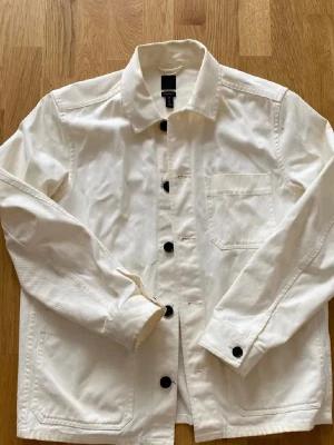 Vit overshirt från H&M - Snygg vit overshirt från H&M med klassisk krage och svarta knappar. Storlek M