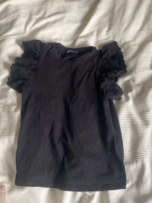 Svart topp med spetsärmar från Zara  - Supersnygg svart topp från Zara med puffiga spetsärmar som ger en extra touch. T-shirten har rund halsringning och är tillverkad i mjukt material. Perfekt för dig som vill ha något stilrent men ändå unikt.