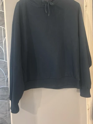 Mörkblå hoodie med huva - En enkel och stilren mörkblå hoodie med huva och dragsko. Hoodien har långa ärmar, ribbade muddar och en avslappnad passform. Perfekt för dig som gillar en clean look och vill ha något bekvämt att slänga på sig. Materialet känns mjukt och skönt mot huden.