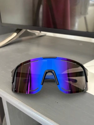 Sportiga blå-lila solglasögon - Säljer ett par sportiga solglasögon med spegelblå och lila tonade linser och svart båge. Sköldformad design som ger en futuristisk look och extra skydd. Perfekta för dig som vill sticka ut och samtidigt få bra skydd mot solen.