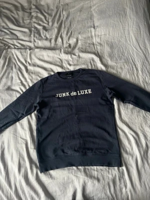 Mörkblå sweatshirt från Junk de Luxe - Är M men sitter som S. Snygg mörkblå sweatshirt från Junk de Luxe med vit logotyptext på bröstet. Tröjan har rund halsringning, långa ärmar och ribbade muddar vid ärmslut och nederkant. Perfekt för en avslappnad och trendig stil.