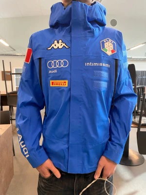 Kappa 6CENTO 611 FISI Italy Ski Team Jacket - Blå vindjacka från Kappa med huva, stora vita ITA-bokstäver på ryggen och flera loggor framtill, bland annat Audi, Pirelli och Intimissimi. Jackan har dragkedjor på sidorna och sportig design, perfekt för dig som gillar italiensk stil. Använd av italienska herr landslaget i skidor. 