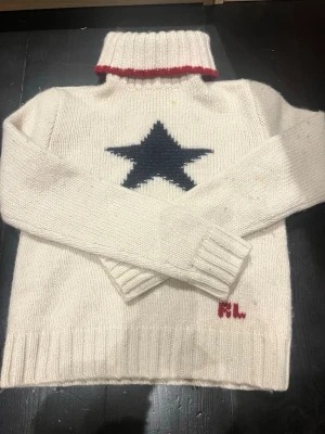 Vit stickad tröja med stjärna Ralph Lauren 2008 - Vit stickad polotröja från Ralph Lauren med en stor mörkblå stjärna på bröstet och röd detalj vid halsen. Tröjan är gjord av ull och silke, med RL-broderi i rött nedtill. Den har några fläcka och lite noppig (se bild) men väldigt snygg! Tröjan är från 2008 och är perfekt för kalla dagar och snygg till jeans. Skriv om du har fler frågor!❤️