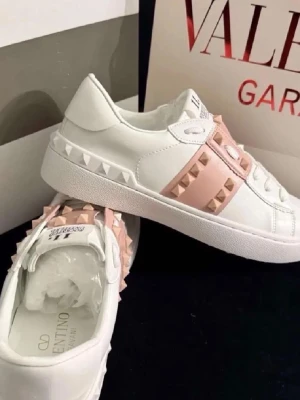 Valentino vita sneakers med nitar - Snygga vita sneakers från Valentino med ljusrosa band och ikoniska nitar i både vitt och rosa. Skorna är låga och har en chunky sula. Tillverkade i läder med coola detaljer som ger en lyxig och trendig vibe.