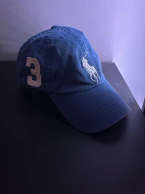 Blå keps från Polo Ralph Lauren - Snygg blå keps från Polo Ralph Lauren med klassisk vit broderad logga framtill och en stor vit siffra 3 på sidan. Kepsen har böjd skärm och justerbar rem bak. Perfekt accessoar för en sportig och stilren look.