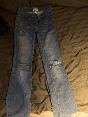Blå bootcut jeans  - Säljer ett par blå bootcut jeans från John Bamer med slitningar på låret och knät. Jeansen har klassisk femficksmodell, låg midja och snygga detaljer på bakfickorna. Perfekta för en avslappnad och trendig look.