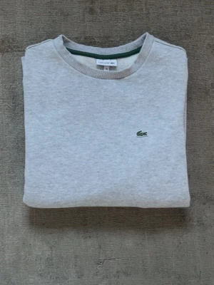 Grå sweatshirt från Lacoste - Klassisk grå sweatshirt från Lacoste med rund halsringning och den ikoniska gröna krokodilloggan broderad på bröstet. Tröjan har ribbade muddar vid ärmslut och nederkant. Perfekt för en avslappnad och stilren look.