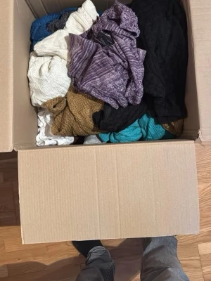 Stor låda med blandade kläder - En stor flyttlåda fylld med olika klädesplagg i flera färger som svart, vitt, lila, blått, turkos och brunt. Plaggen verkar vara av stickat och bomullsmaterial och innehåller både tunna och tjockare tröjor samt toppar. Perfekt för dig som vill ha variation i garderoben.